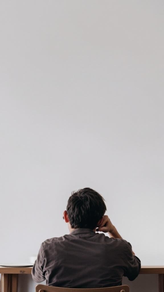 A person sitting at a table, looking contemplative with a blank wall in front of them.