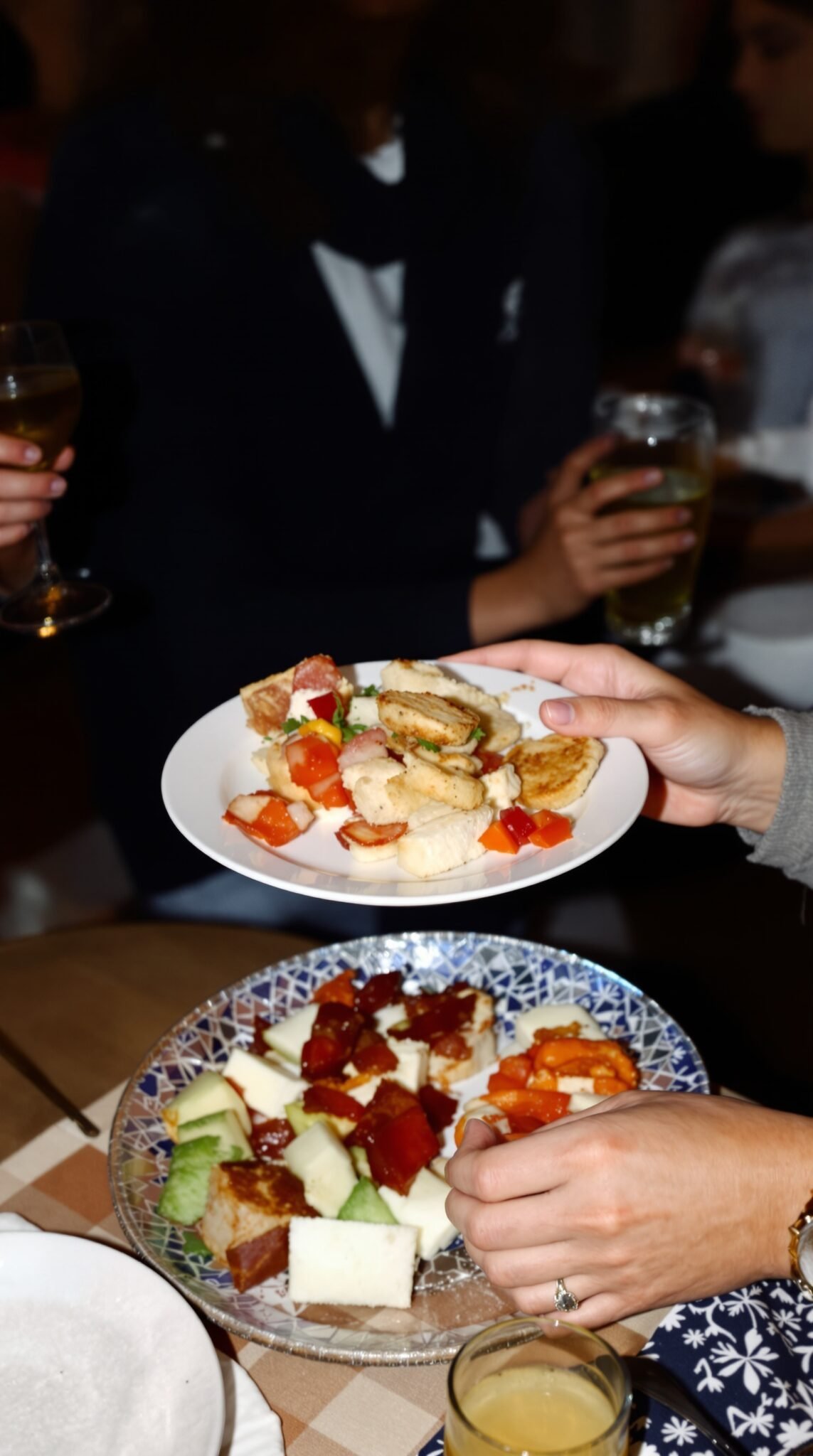 A hand holding a plate of food at a social gathering, surrounded by friends enjoying drinks and conversation.
